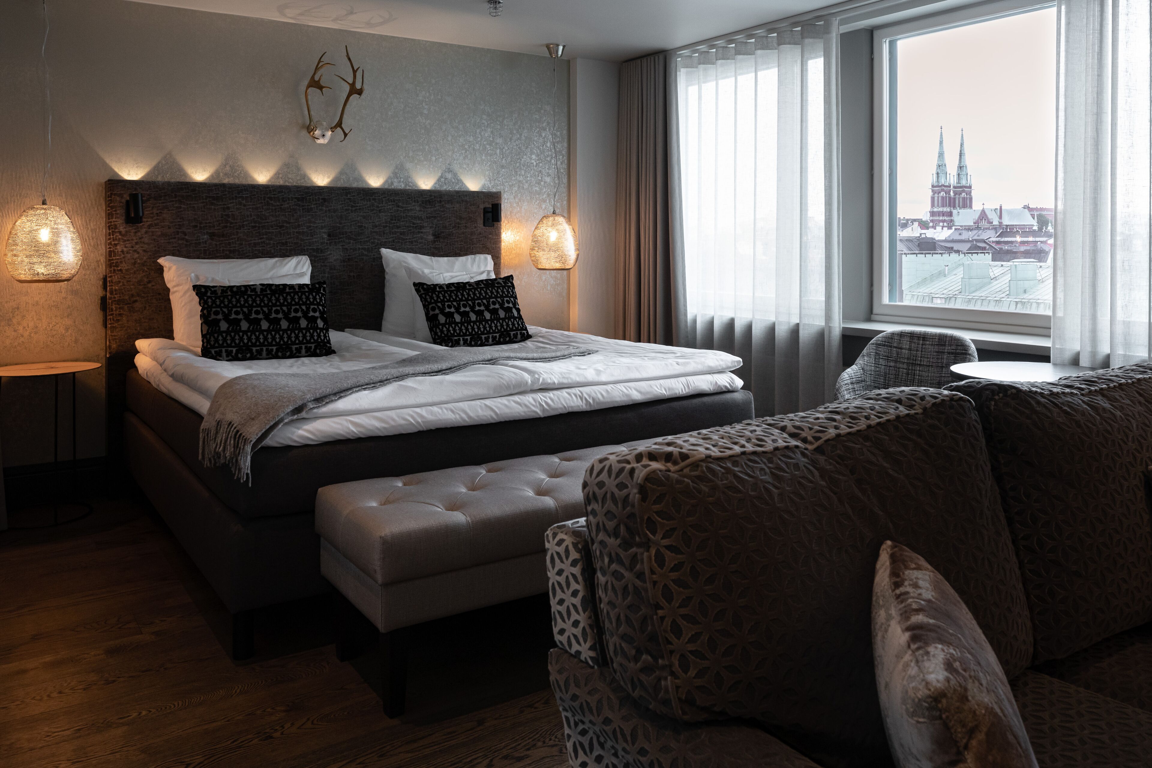Ein modernes Hotelzimmer mit einem großen Bett, einer Couch, Blick auf eine Stadtlandschaft und einer Wand mit Geweihen.
