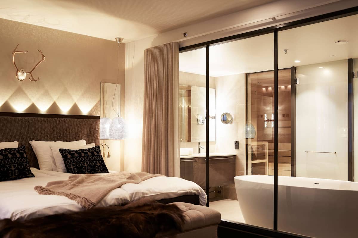 A modern hotel room with a large bed, a glass-enclosed bathroom, and a view of the interior.