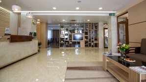 Área de recepção de hotel com piso de mármore, balcão de atendimento, televisão e um espaço de estar com um vaso de flores.