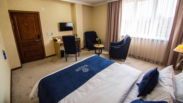 Quarto de hotel com uma cama, uma escrivaninha, uma cadeira, uma televisão e janela com cortinas.