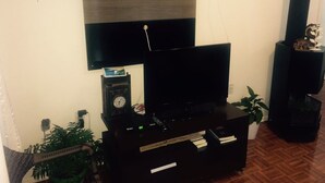 Ein Wohnzimmer mit einem Flachbildfernseher auf einem schwarzen Standfuß, einer Uhr und gerahmten Bildern an der Wand.