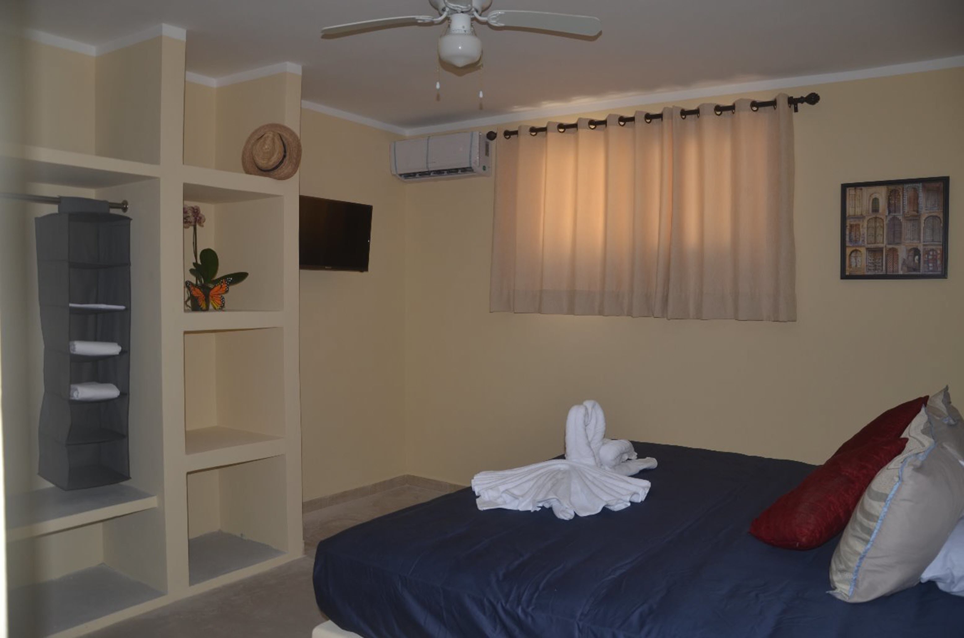 A hotel room with a bed, a ceiling fan, a window with curtains, a wall-mounted air conditioner, and a shelf with a plant.