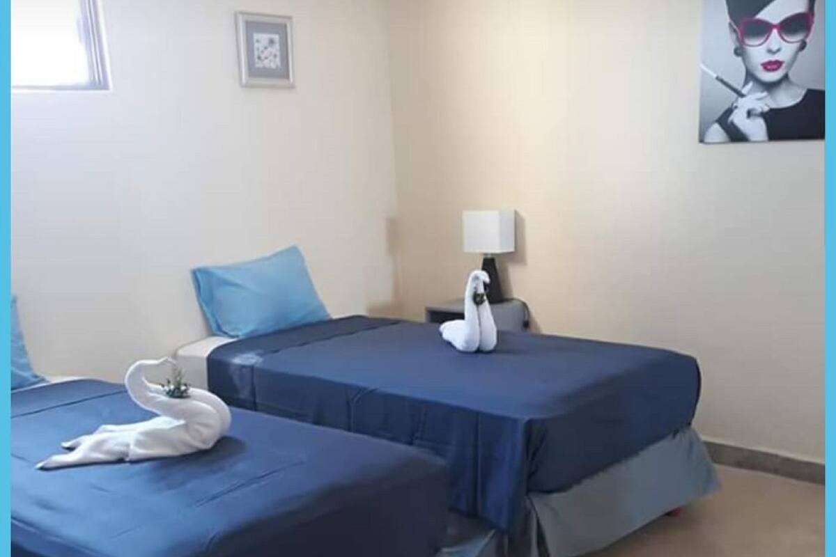 Habitación de hotel con dos camas, cada una con cubrecama azul y almohada con forma de cisne blanco. Hay una mesita de noche con una lámpara y un cuadro en la pared.