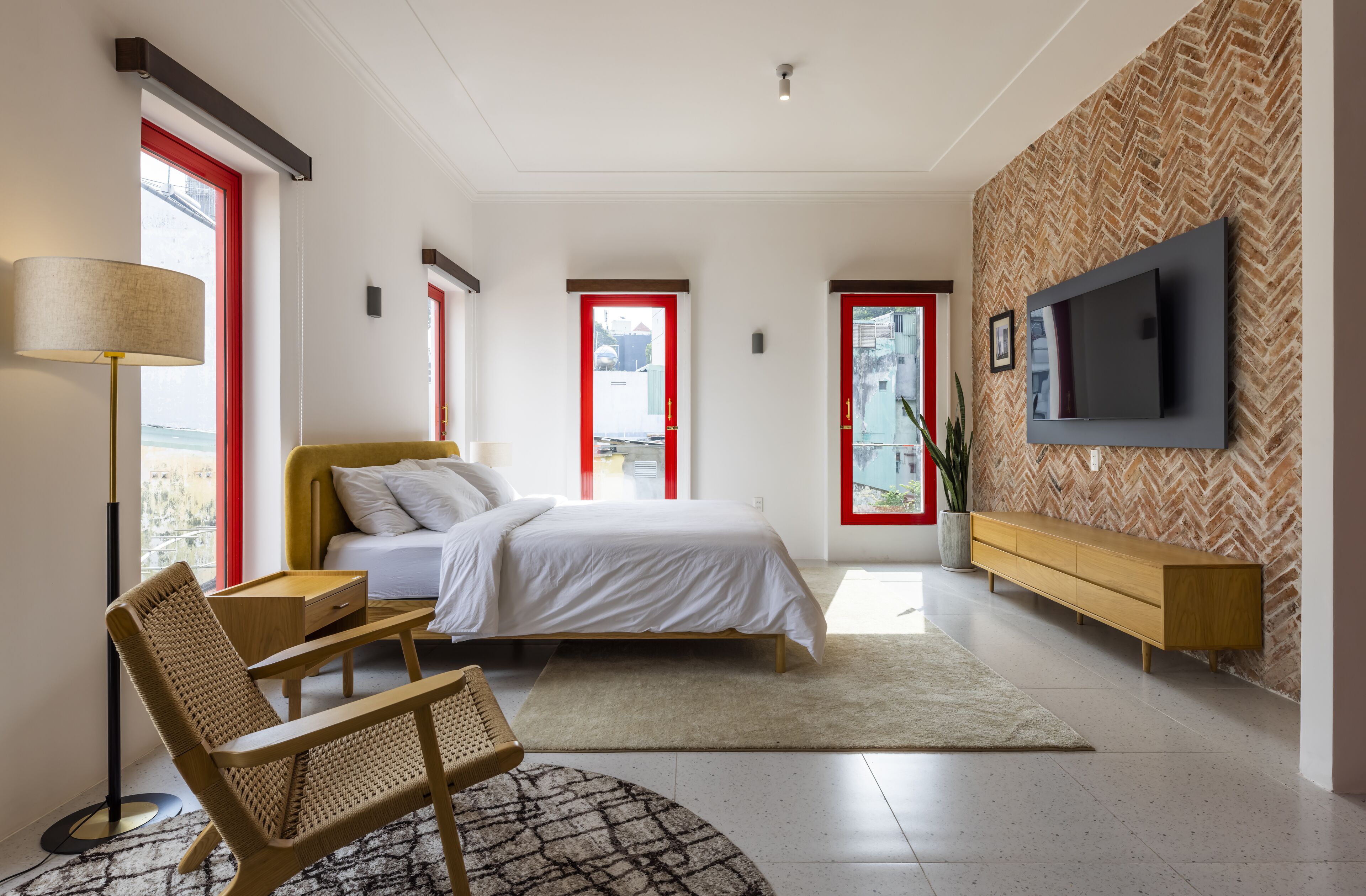 A modern bedroom with a bed, a wooden bench, a TV mounted on a herringbone-patterned wall, and a large window with red frames.