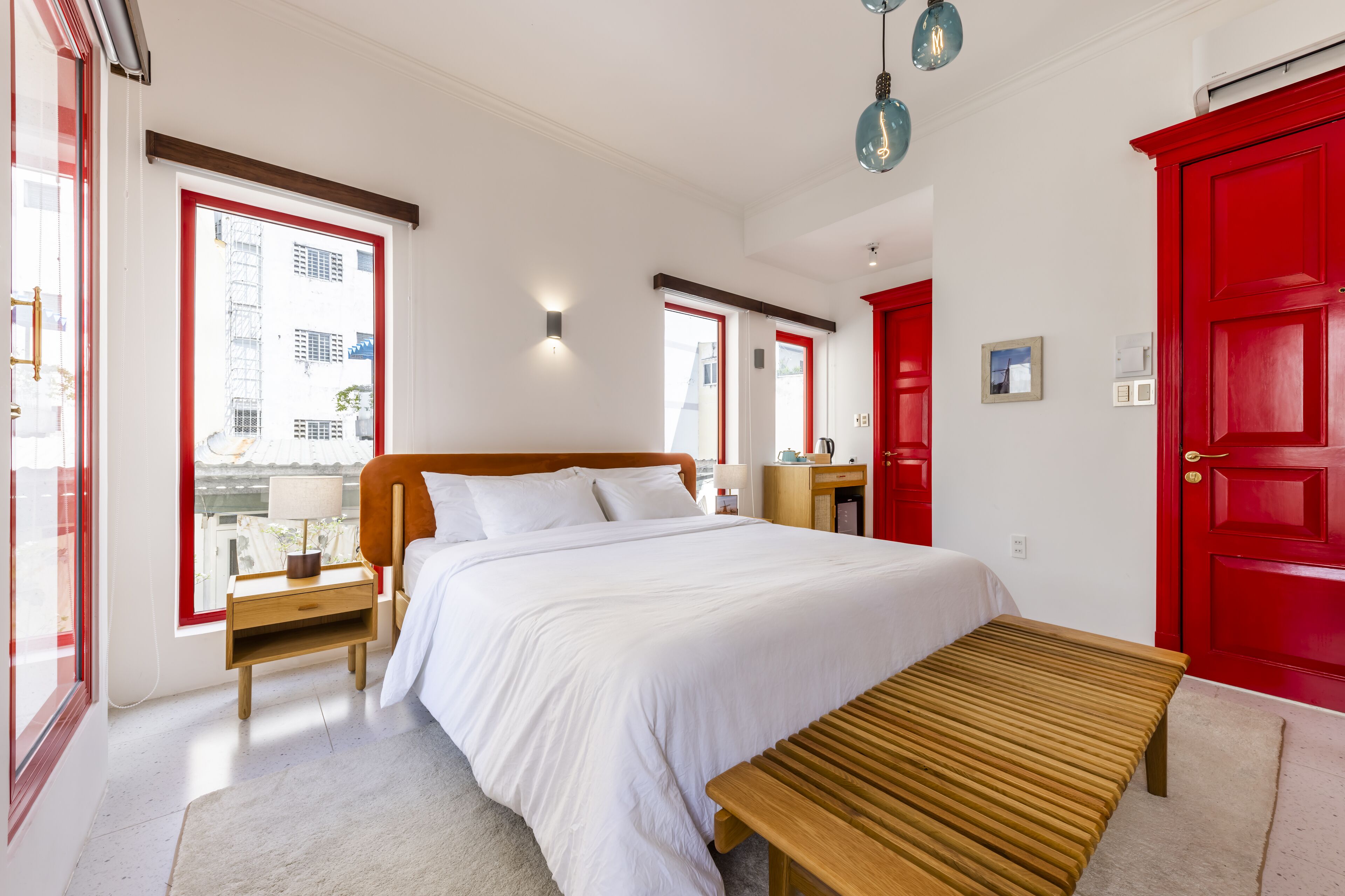 Un dormitorio con una cama grande, un banco de madera, una puerta roja y vistas de edificios a través de la ventana.