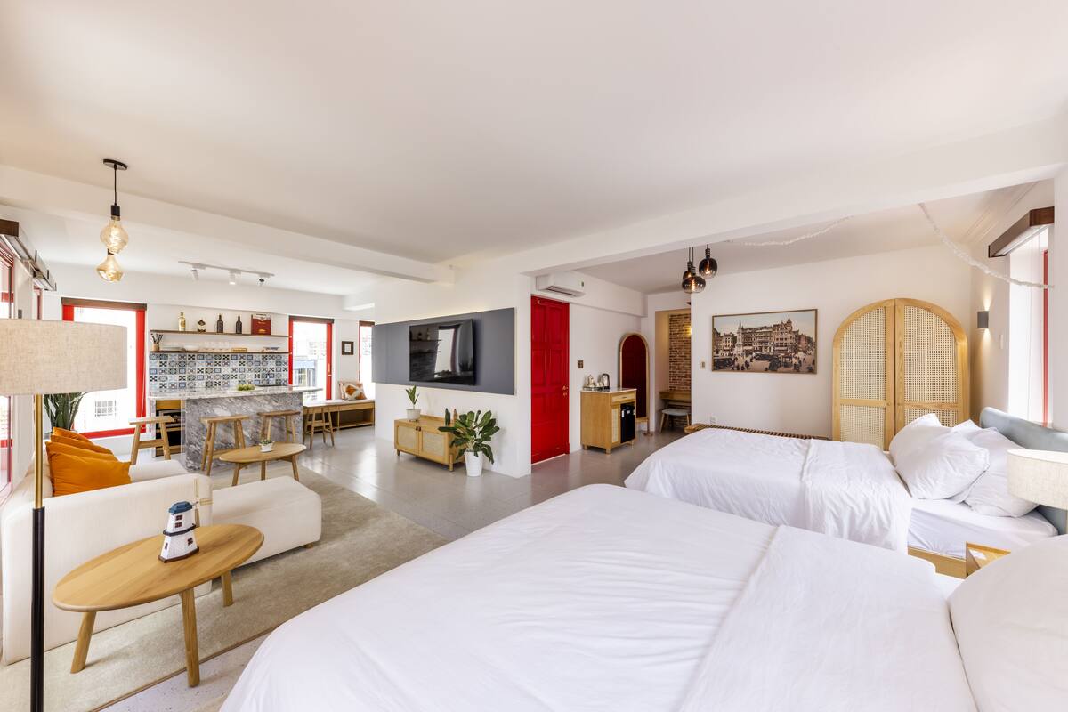 Una habitación de hotel moderna con dos camas, una zona de estar con un sofá y una mesita, una cocina con barra y un comedor.