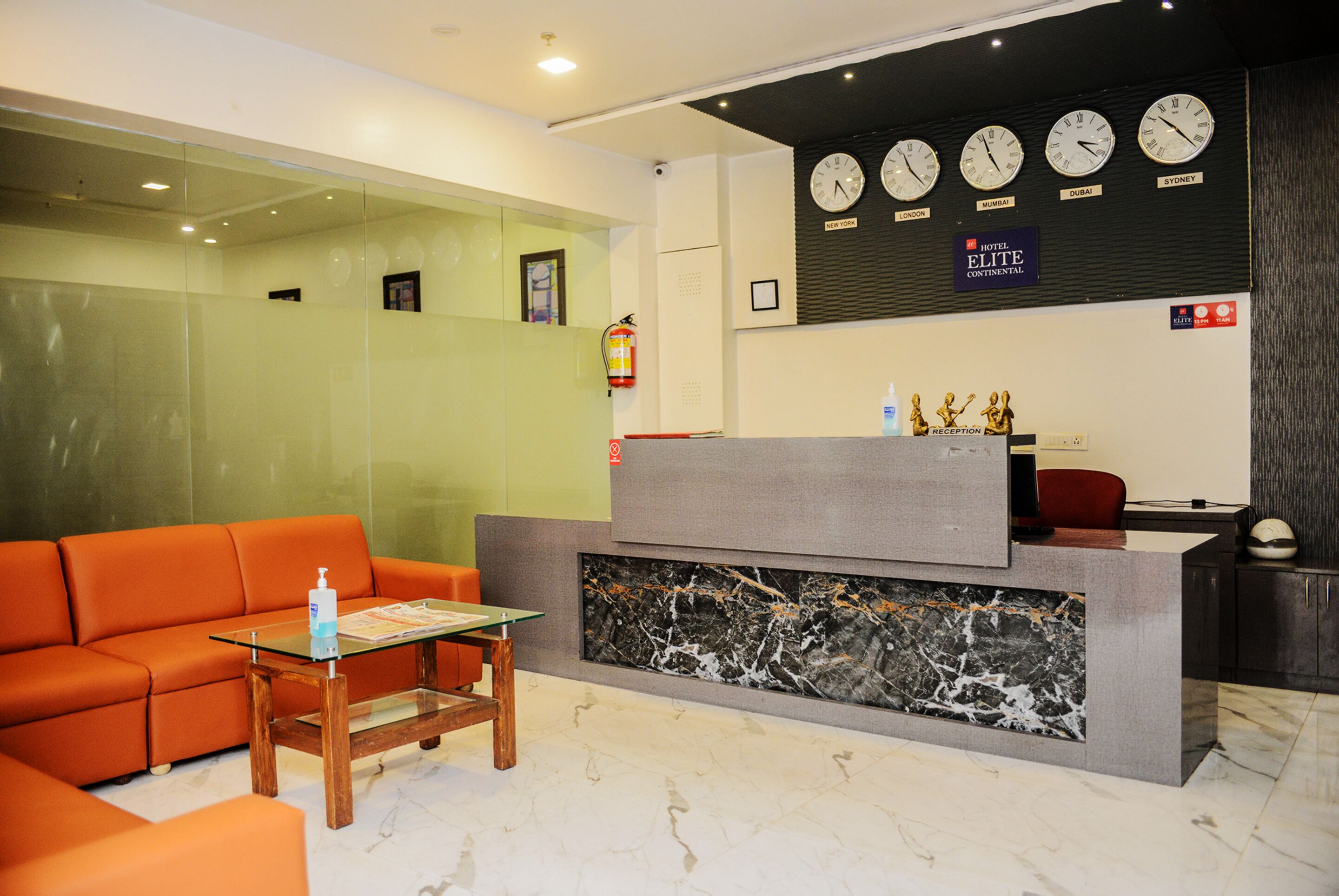A hotel reception area with a reception desk, a glass-top table, an orange sofa, and a marble wall.