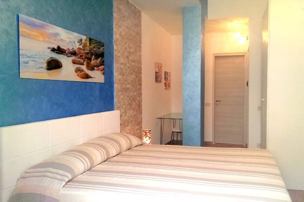 Un dormitorio con cama, un cuadro de pájaros en la pared, una mesita y una puerta que da a otra habitación.
