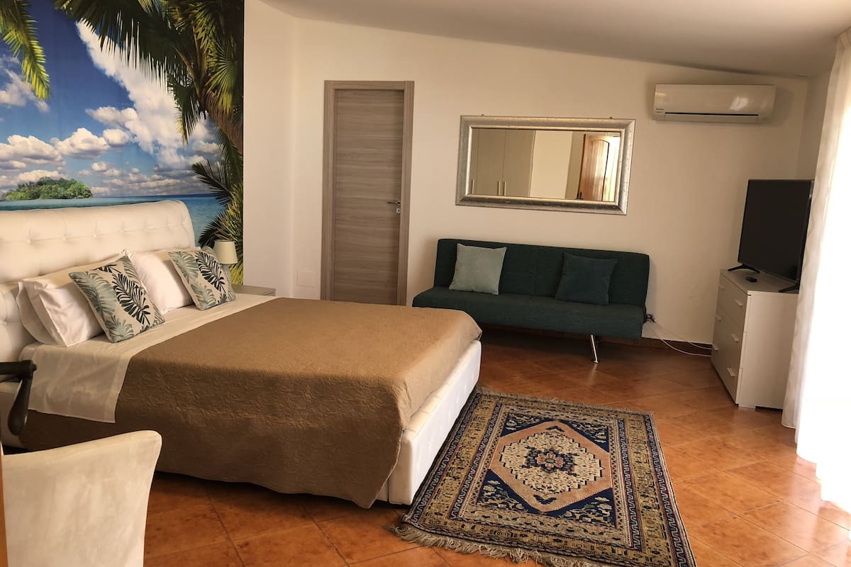 Un dormitorio con cama, sofá, televisor y un cuadro de paisaje tropical.
