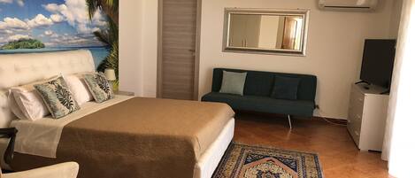 A bedroom with a bed, a sofa, a TV, and a painting of a tropical scene.