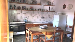 A kitchen with a stove, oven, refrigerator, and a dining table with chairs. There are shelves above the counter with various kitchen items and a clock on the wall. - I Faraglioni (Terrasini)