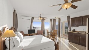 A hotel room with a bed, a desk, a chair, a ceiling fan, and a kitchen area. - Turtles Nest Beach Resort  (West End Village)