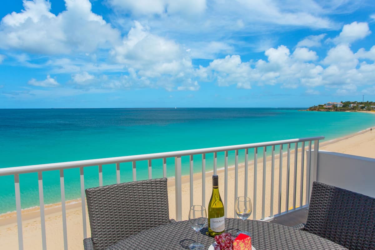Un balcon agrémenté de meubles en osier, une bouteille de vin et des verres posés sur une table, le tout donnant sur une plage aux eaux turquoise et limpides, avec une balustrade blanche.