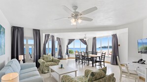 A spacious living room with a ceiling fan, a dining area with a table and chairs, and large windows offering a view of the sea. - Turtles Nest Beach Resort  (West End Village)
