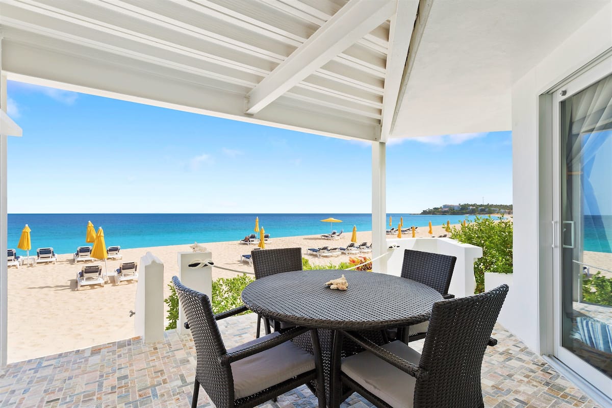 Un espace extérieur abrité, équipé d’une table et de chaises, donnant sur une plage parsemée de parasols et de chaises longues.