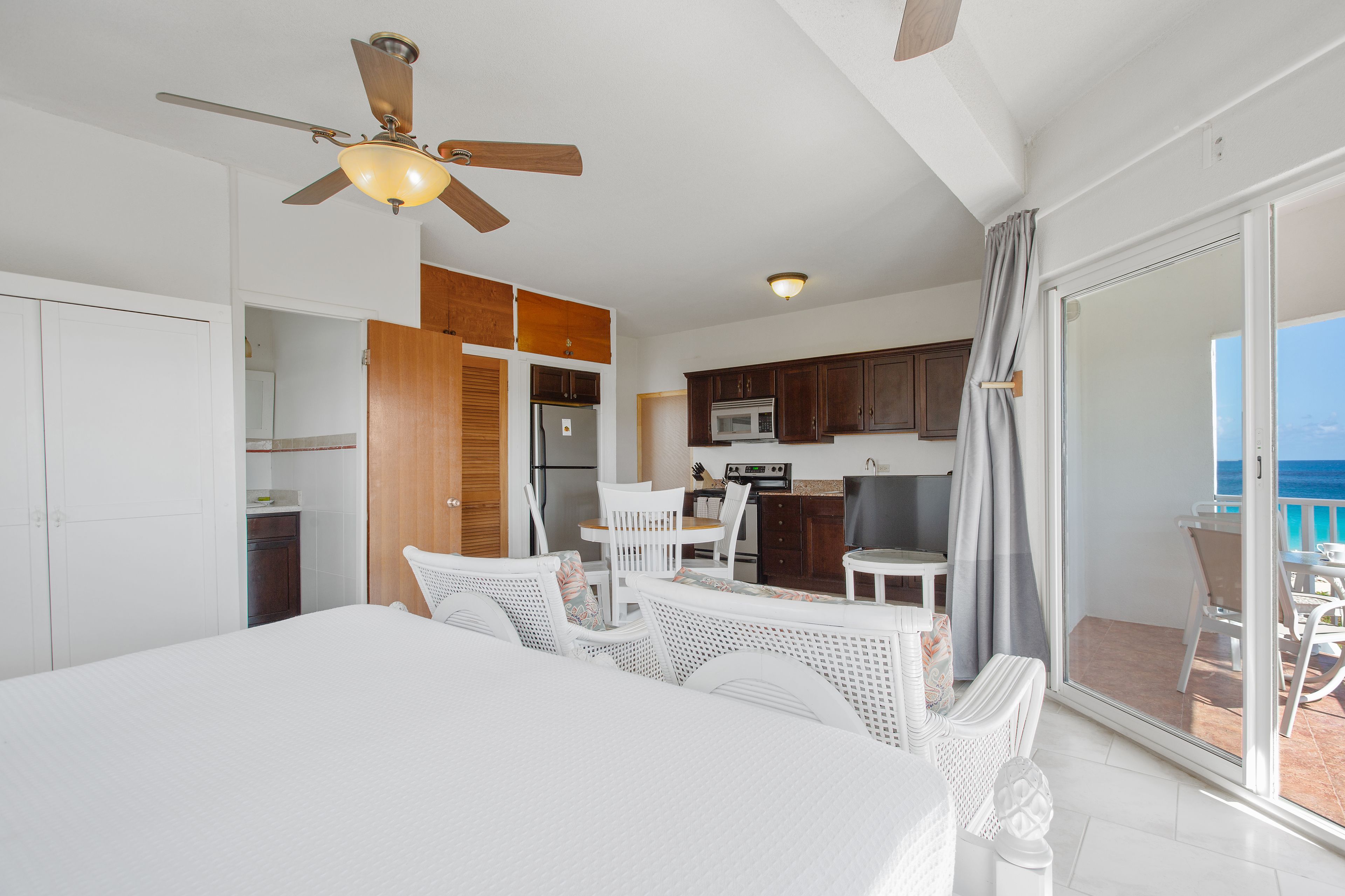Habitación de hotel con balcón, ventilador de techo, kitchenette y zona de comedor con mobiliario blanco.