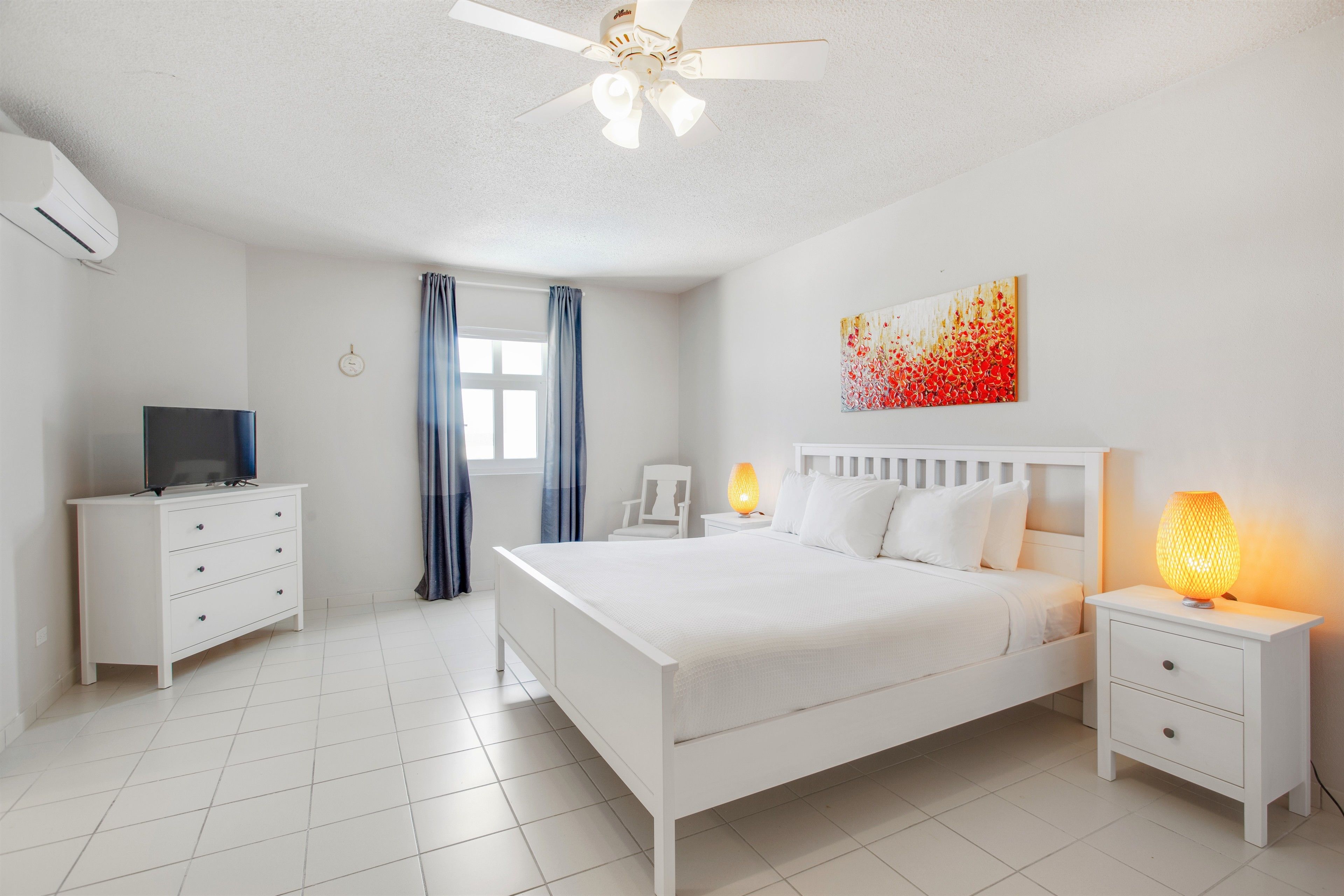 Ein helles Schlafzimmer mit Bett, Fernseher, Nachttisch und Deckenventilator.