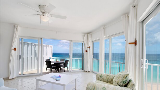 A spacious balcony with a view of the ocean, a white ceiling fan, a dining table with chairs, and a patterned sofa. - Turtles Nest Beach Resort  (West End Village)