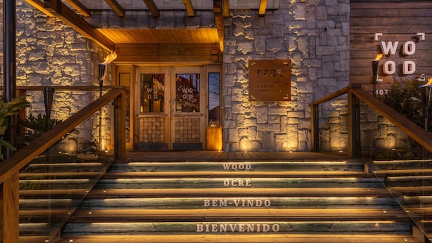 A wooden staircase leading to a stone building entrance with a sign reading "WOOD OCRE BEM-VINDO BIENVENIDO". - Wood Hotel Gramado (Gramado)