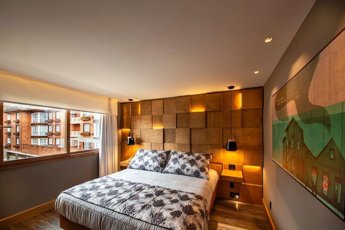 A modern bedroom with a large bed, wooden headboard, two bedside lamps, a view of buildings through a window, and a painting on the wall.