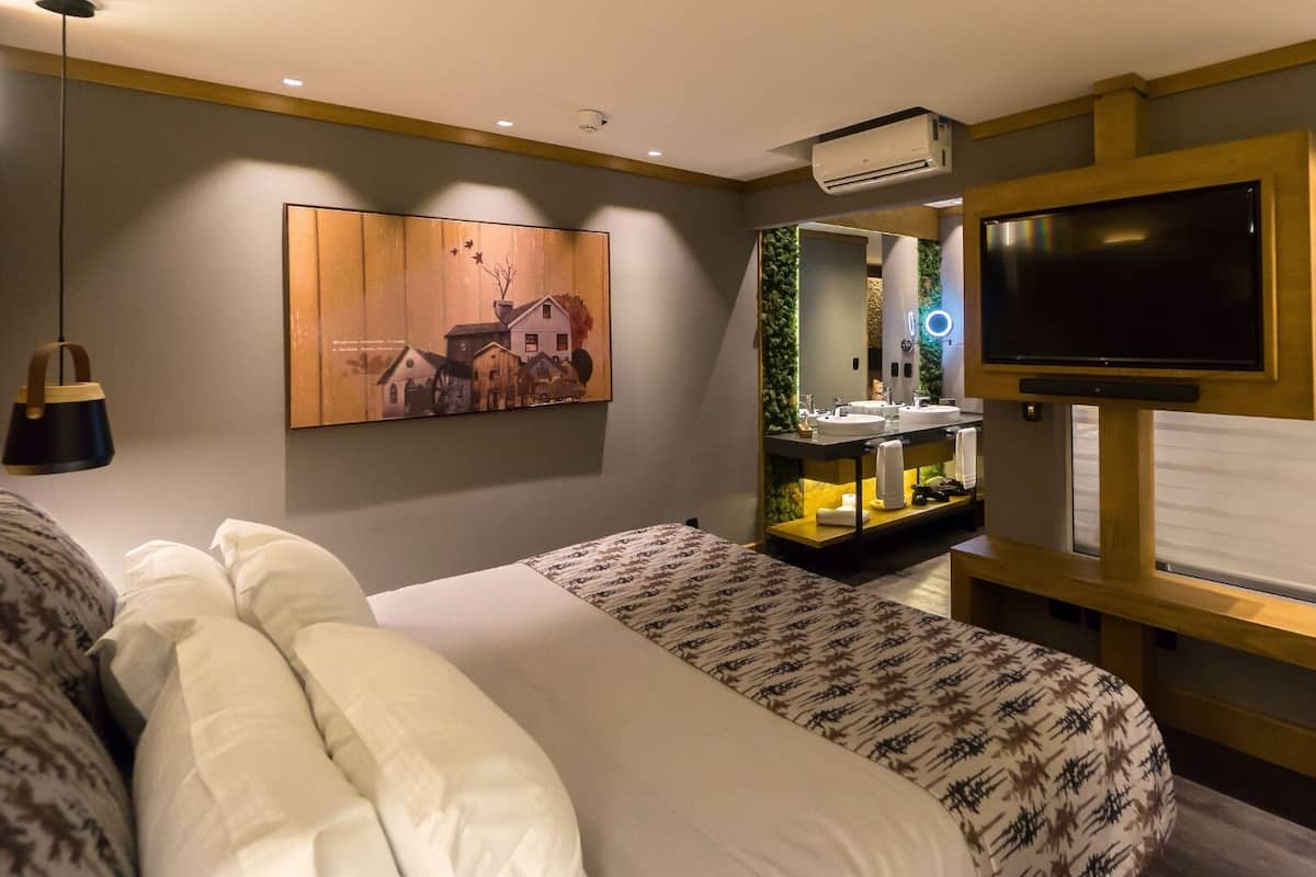 A hotel room with a bed, a TV, a mirror, and a bathroom vanity.