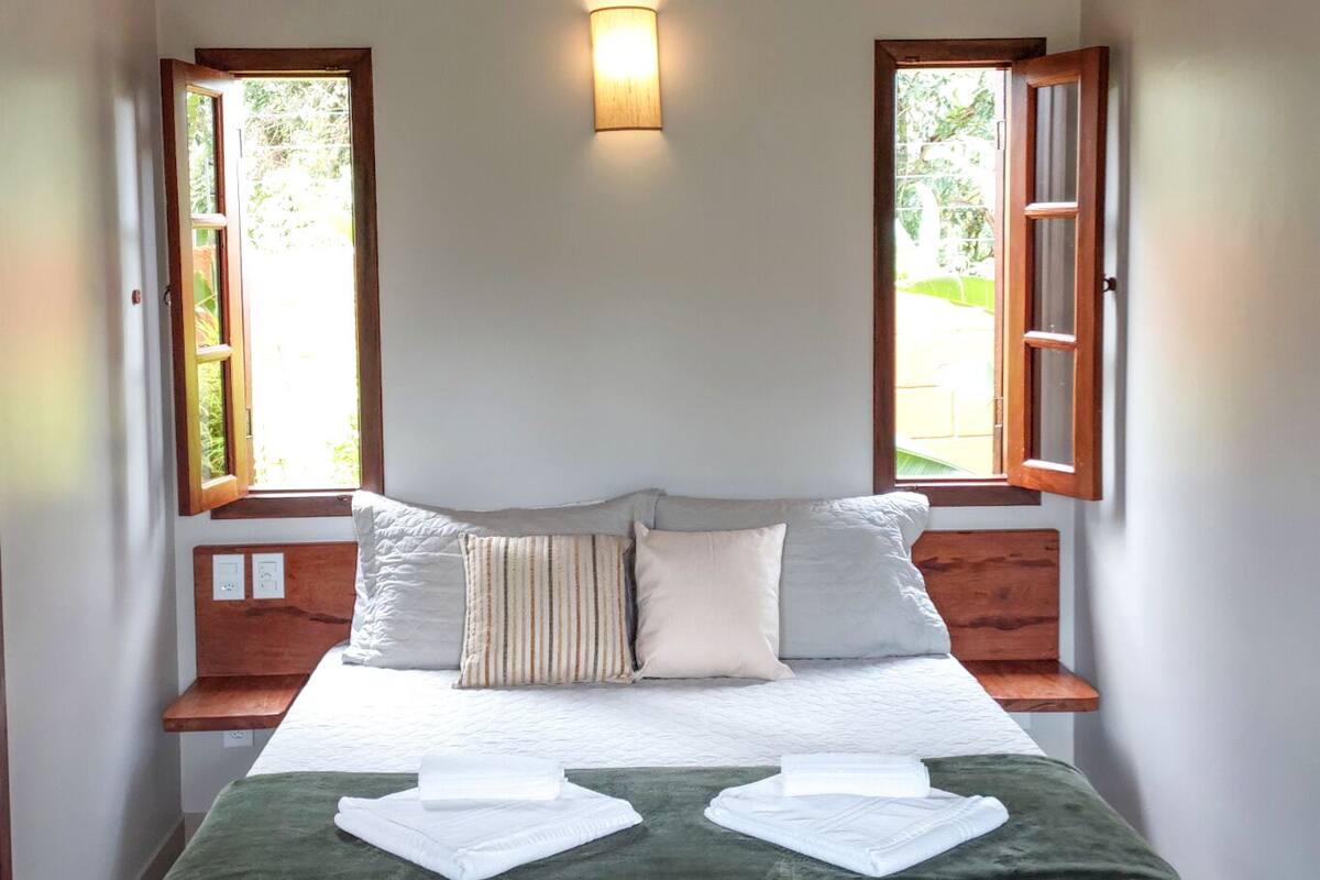 A neatly made bed with pillows and a green throw blanket, two windows with wooden shutters, and a wall-mounted light fixture.