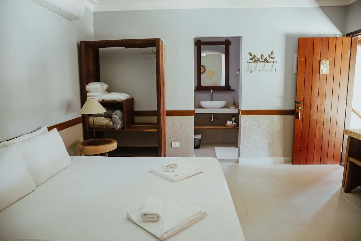 A hotel room with a large bed, a wooden wardrobe, a bathroom with a sink and mirror, and a wooden door.