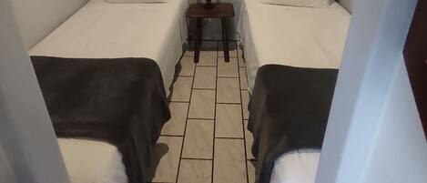 A small, compact room with two single beds, a small table with a lamp, and a window with a blind.