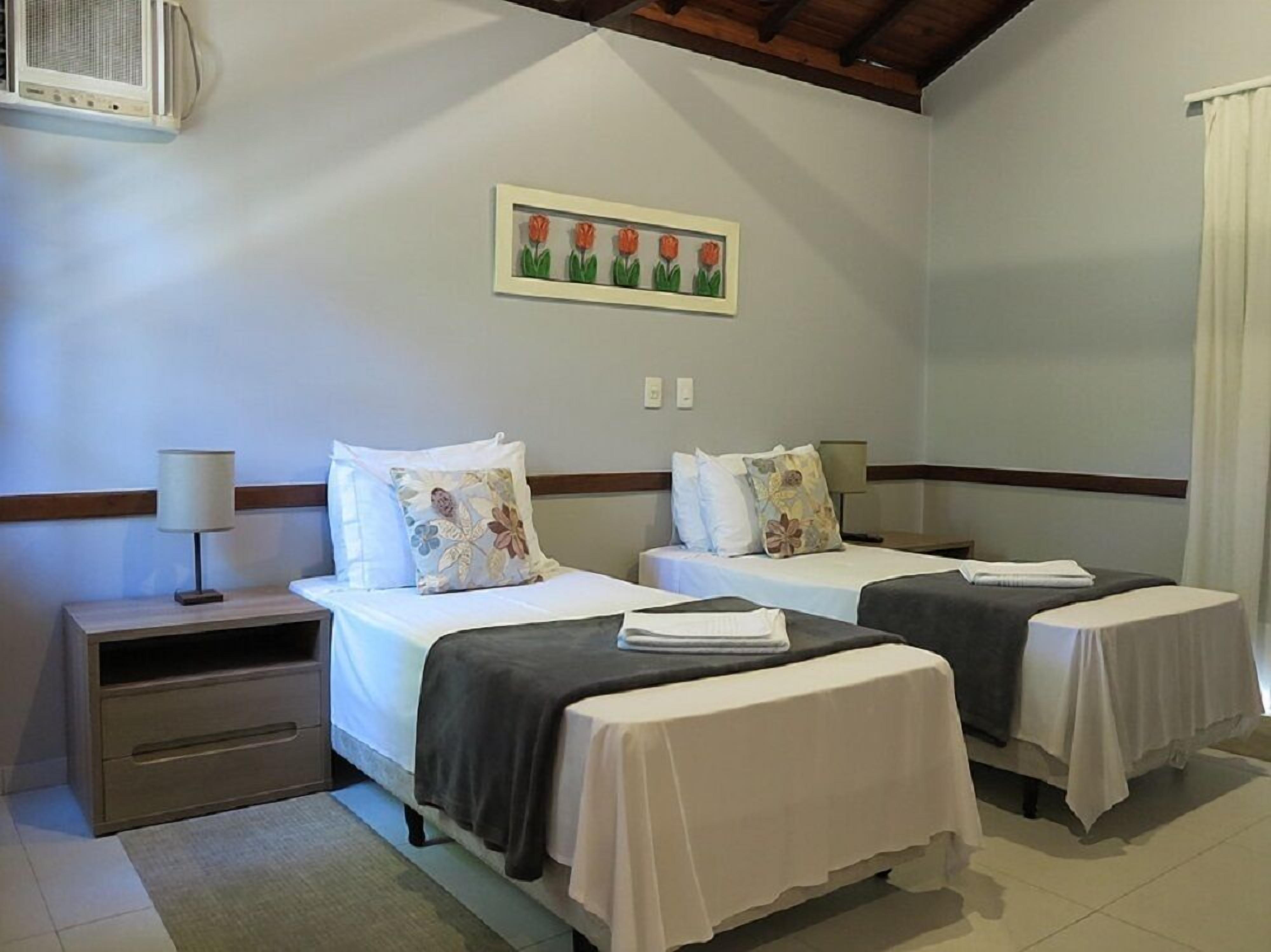 A hotel room with two beds, a nightstand, a lamp, and a painting of flowers on the wall.