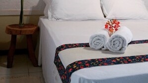 Una cama bien hecha con sábanas blancas, un taburete de madera con una planta y una lámpara fijada a la pared.