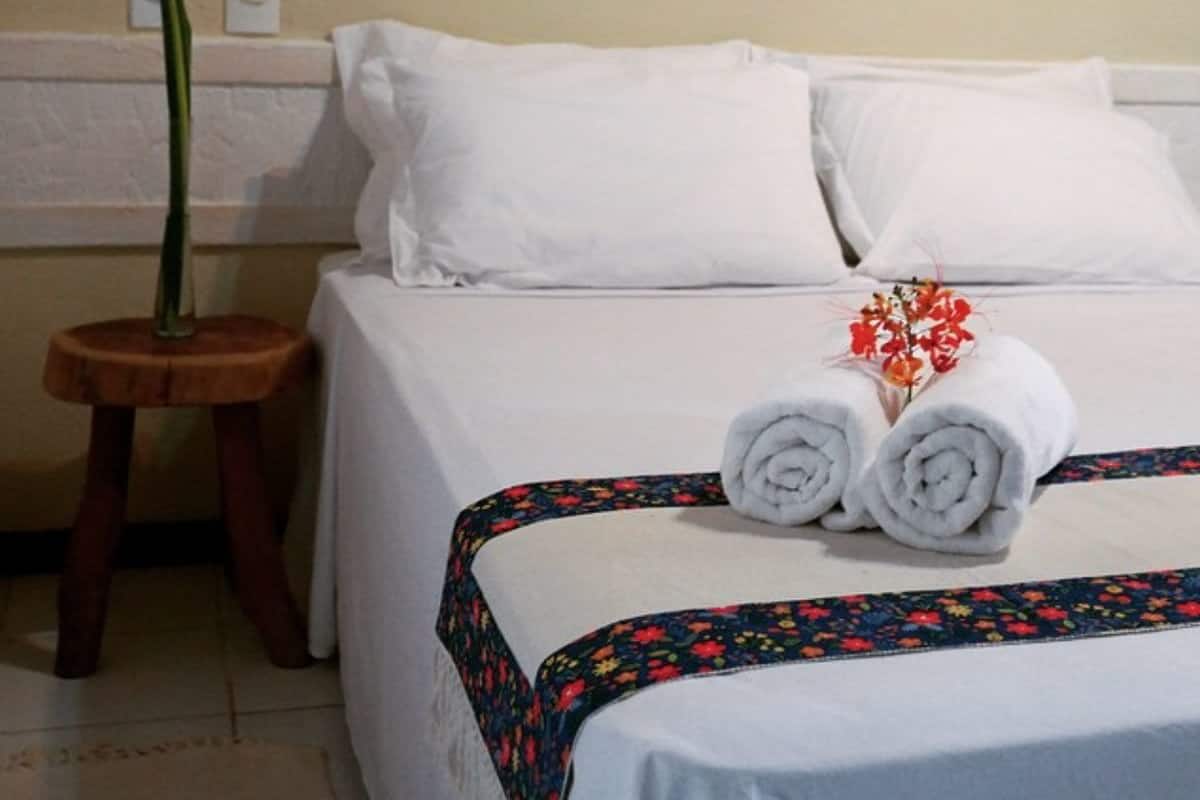 Una cama bien hecha con sábanas blancas, un taburete de madera con una planta y una lámpara fijada a la pared.