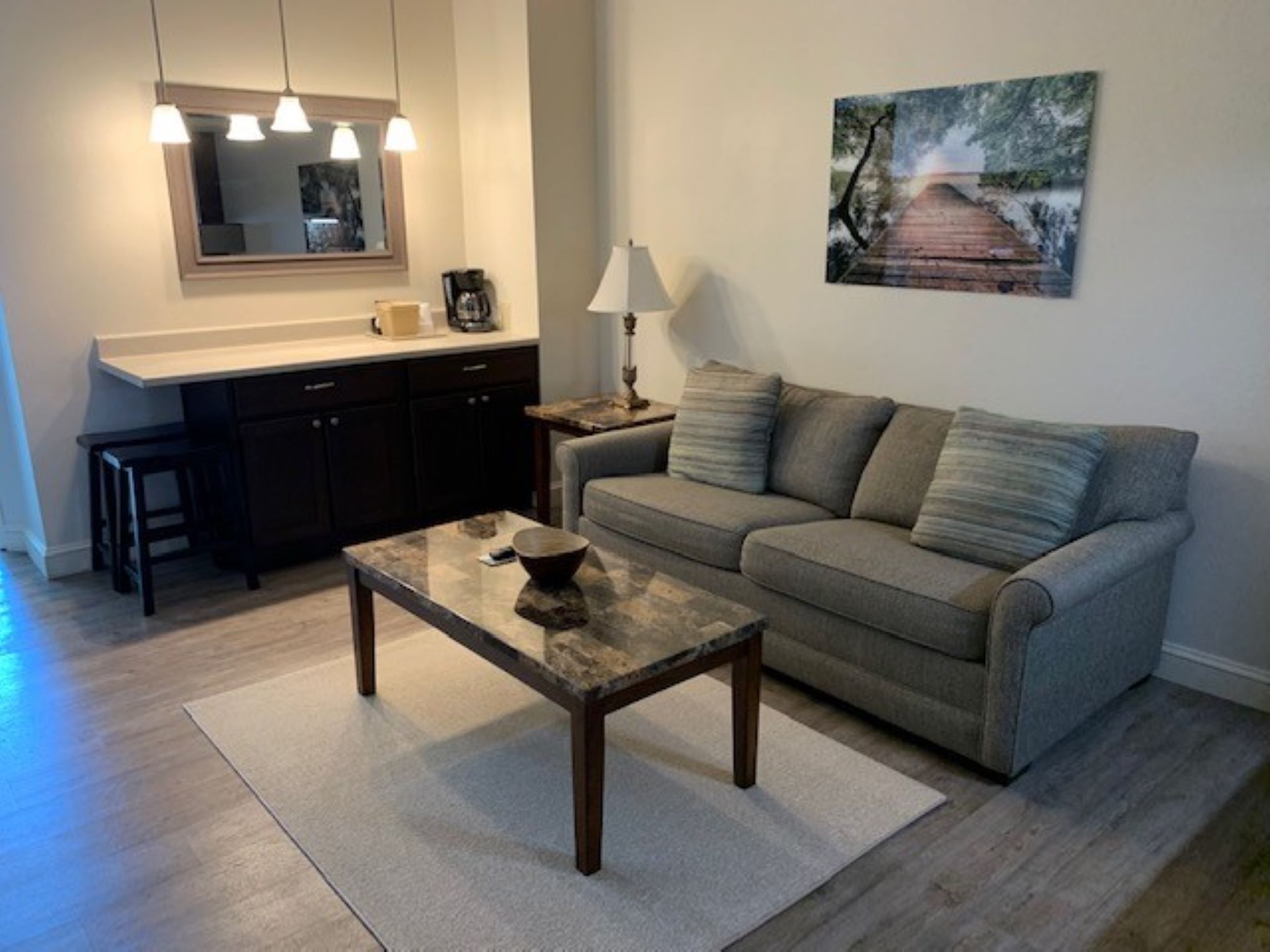 A living room with a sofa, a coffee table, a kitchen area with a counter and cabinets, and a painting on the wall.