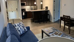 A modern living room with a sofa, a coffee table, and a dining area with a table and chairs. The kitchen is visible with dark cabinets and a microwave. - McCoy's Inn (Ripley)