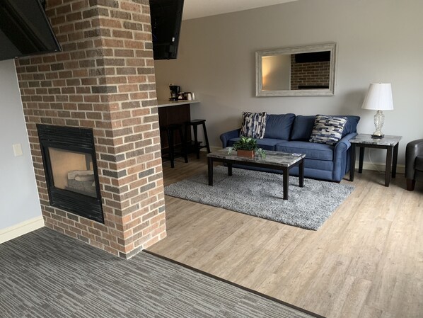 A living room with a brick fireplace, a blue sofa, a coffee table, and a small side table with a lamp.