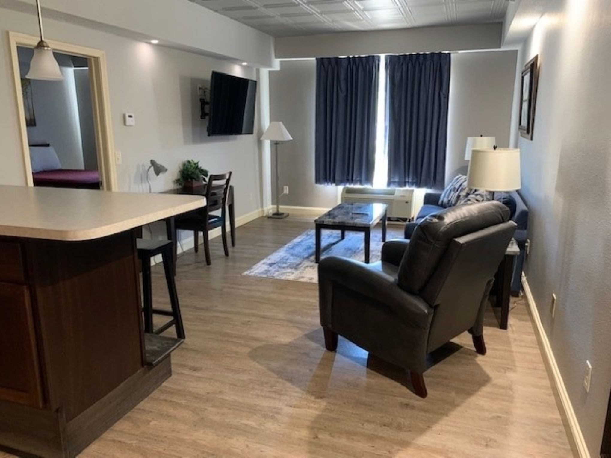 A modern living room with a sofa, armchair, and coffee table. There is a TV mounted on the wall, a window with curtains, and a small table with a lamp.