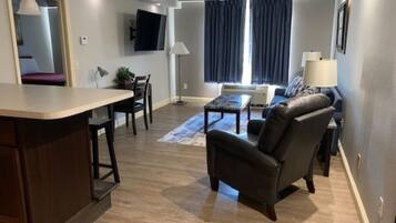 A modern living room with a sofa, armchair, and coffee table. There is a TV mounted on the wall, a window with curtains, and a small table with a lamp.