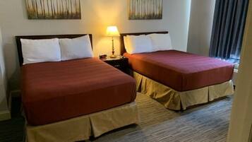 A hotel room with two beds, each with a red bedspread and white pillows. There are two wall-mounted paintings of trees, a bedside lamp, and a window with curtains.