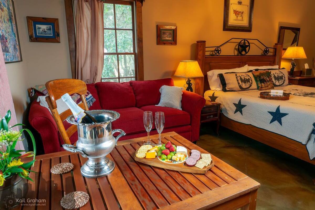 Une chambre d’hôtel chaleureuse avec un canapé rouge, une table en bois sur laquelle sont posés un pichet en argent et un plateau de fruits, un lit recouvert d’une couverture à motifs d’étoiles et une table de chevet avec une lampe.