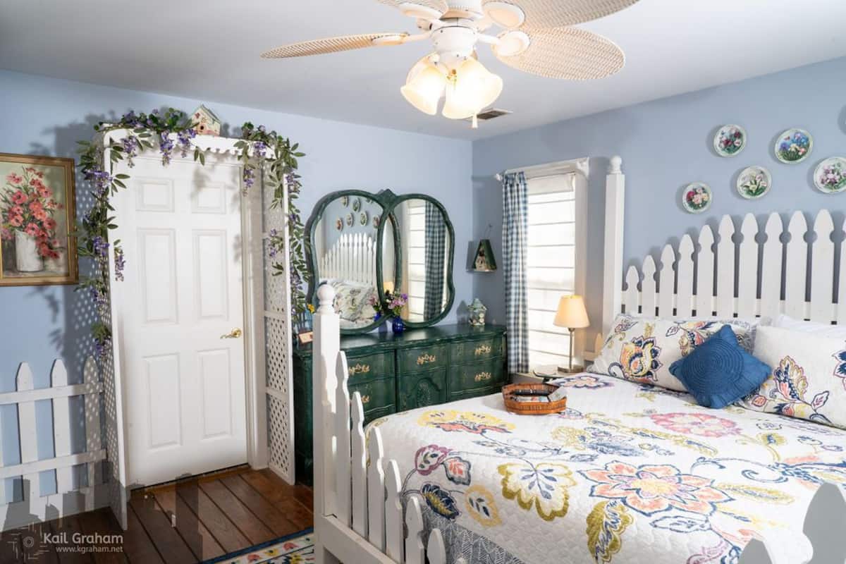 Une chambre à coucher avec un lit blanc, une literie à motifs floraux, une commode verte, une porte blanche et un ventilateur de plafond.