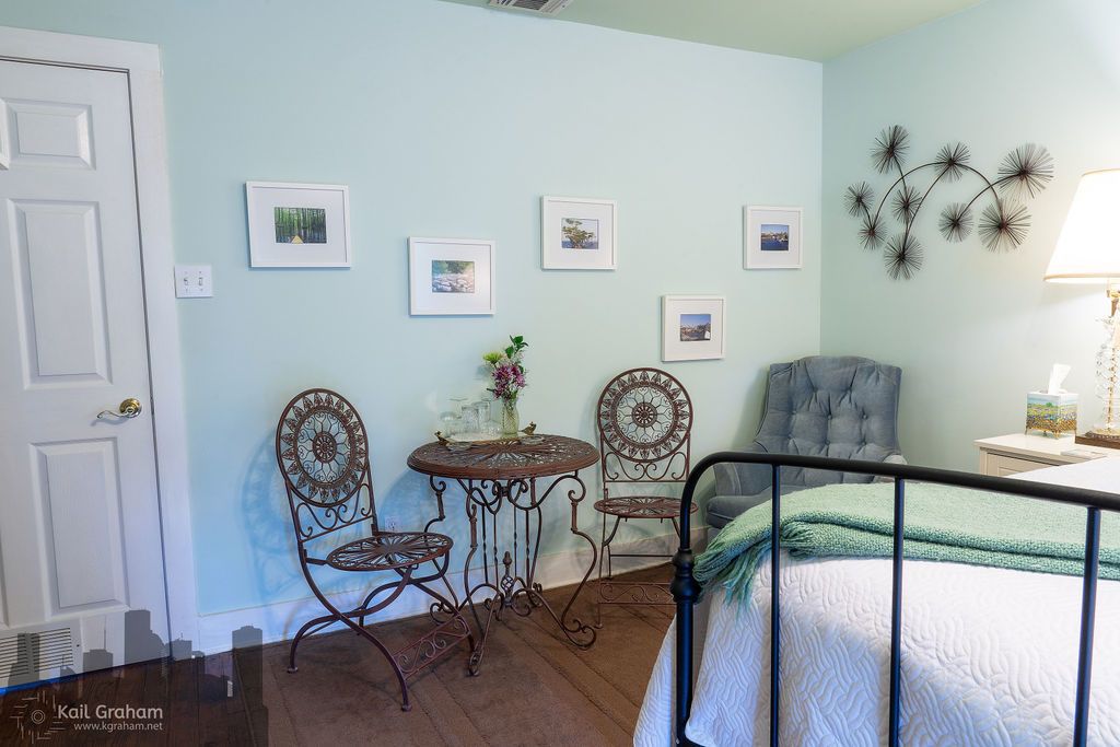A bedroom with a bed, two chairs, a small table with a vase, and wall-mounted pictures.