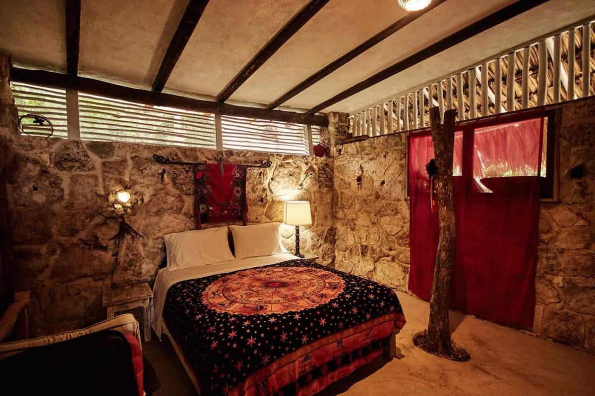 A rustic bedroom with a stone wall, a wooden ceiling, a bed with a patterned bedspread, a nightstand, and a red curtain.