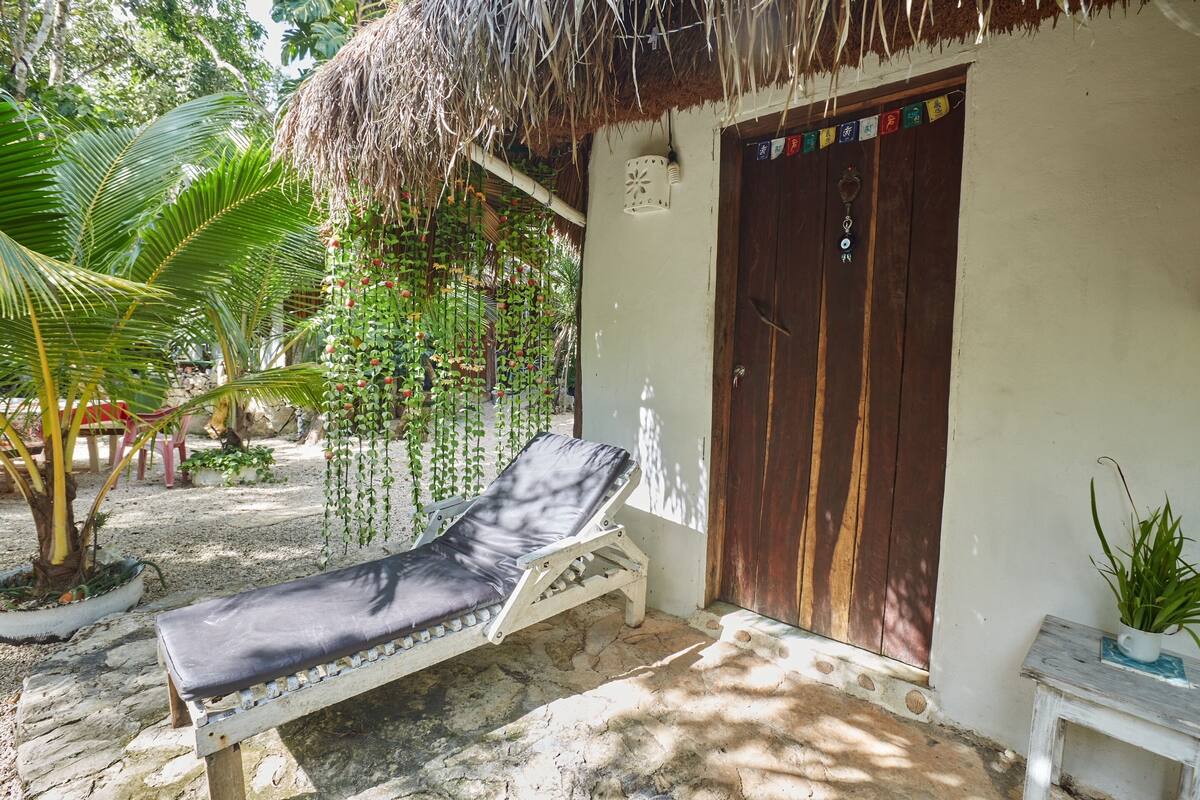 A thatched-roof building with a wooden door, a lounge chair, and hanging plants.