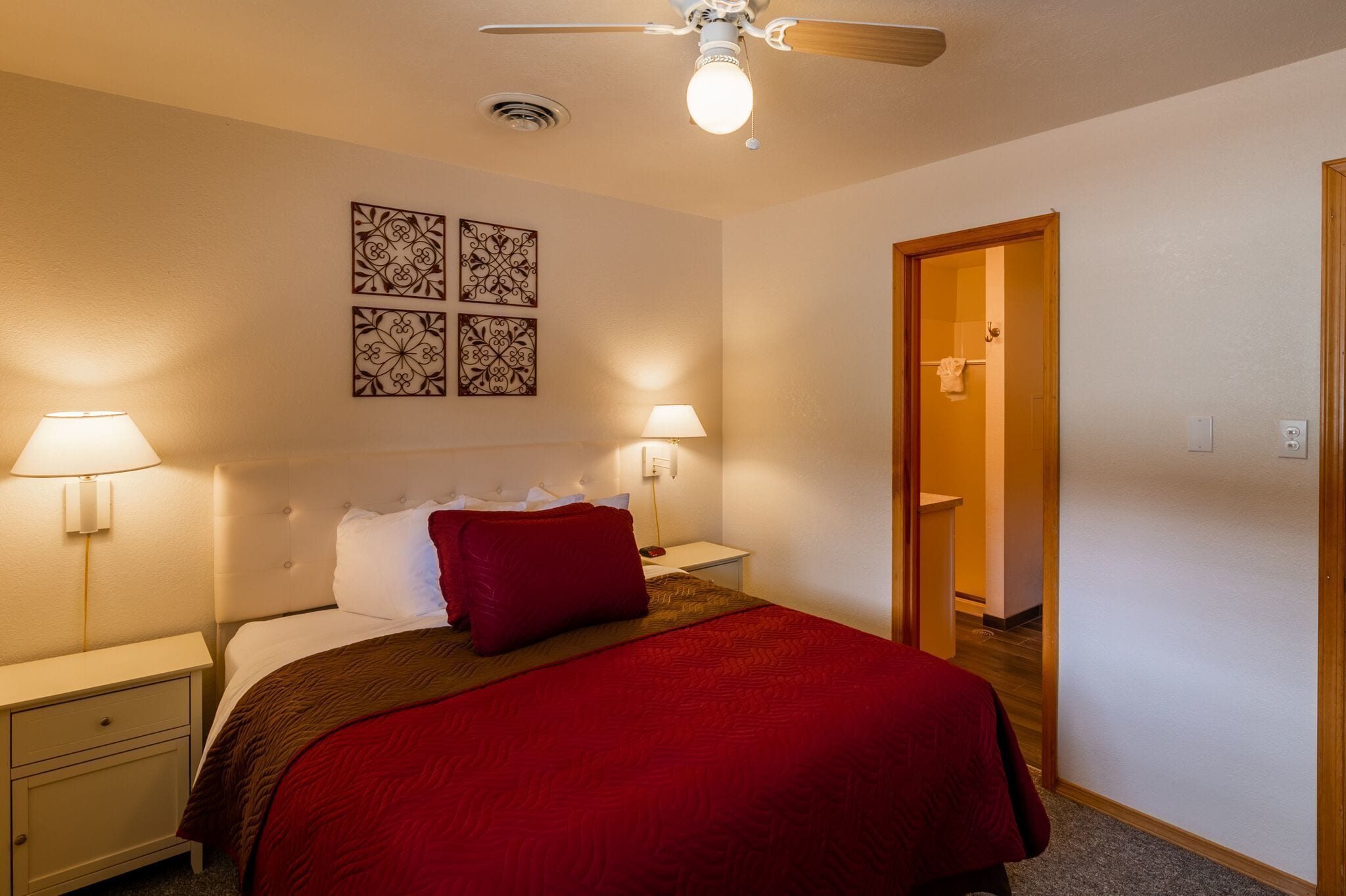A hotel room with a bed, two bedside lamps, a ceiling fan, and two wall-mounted decorative pieces.