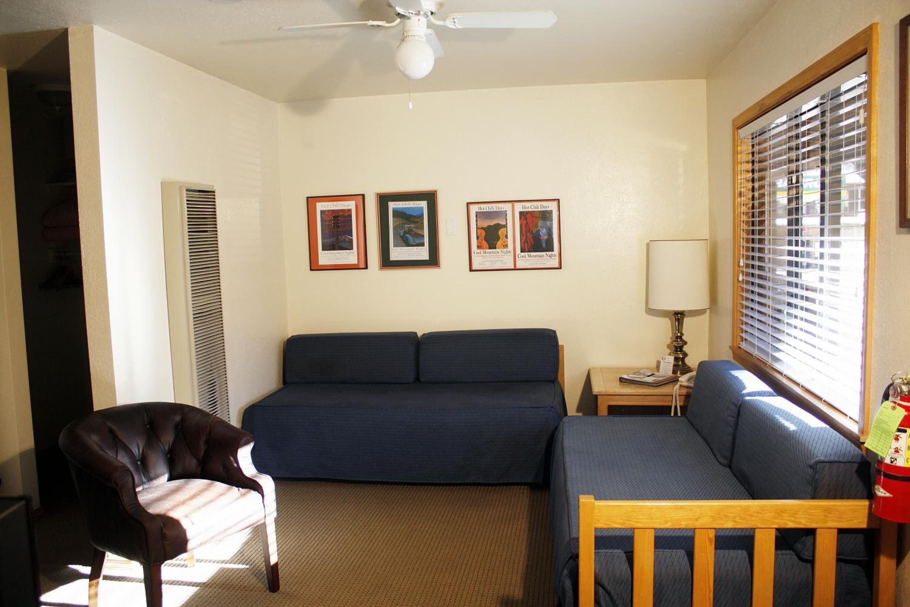 A room with a bed, a chair, and a small table with a lamp. There are three framed pictures on the wall.