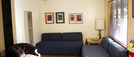 A room with a bed, a chair, and a small table with a lamp. There are three framed pictures on the wall.
