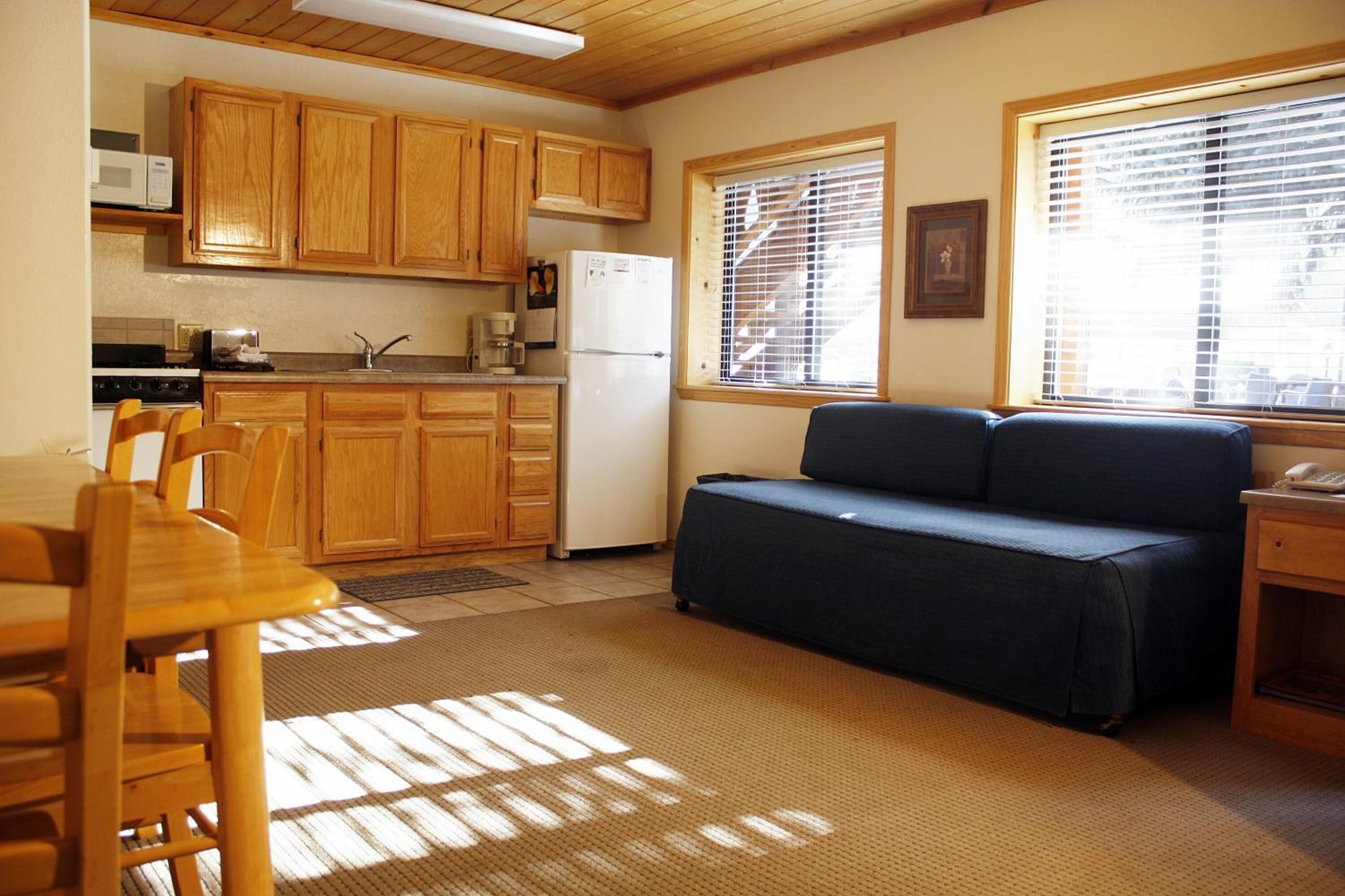 A compact kitchen with wooden cabinets, a microwave, and a refrigerator. There is a dining table with chairs and a blue sofa.