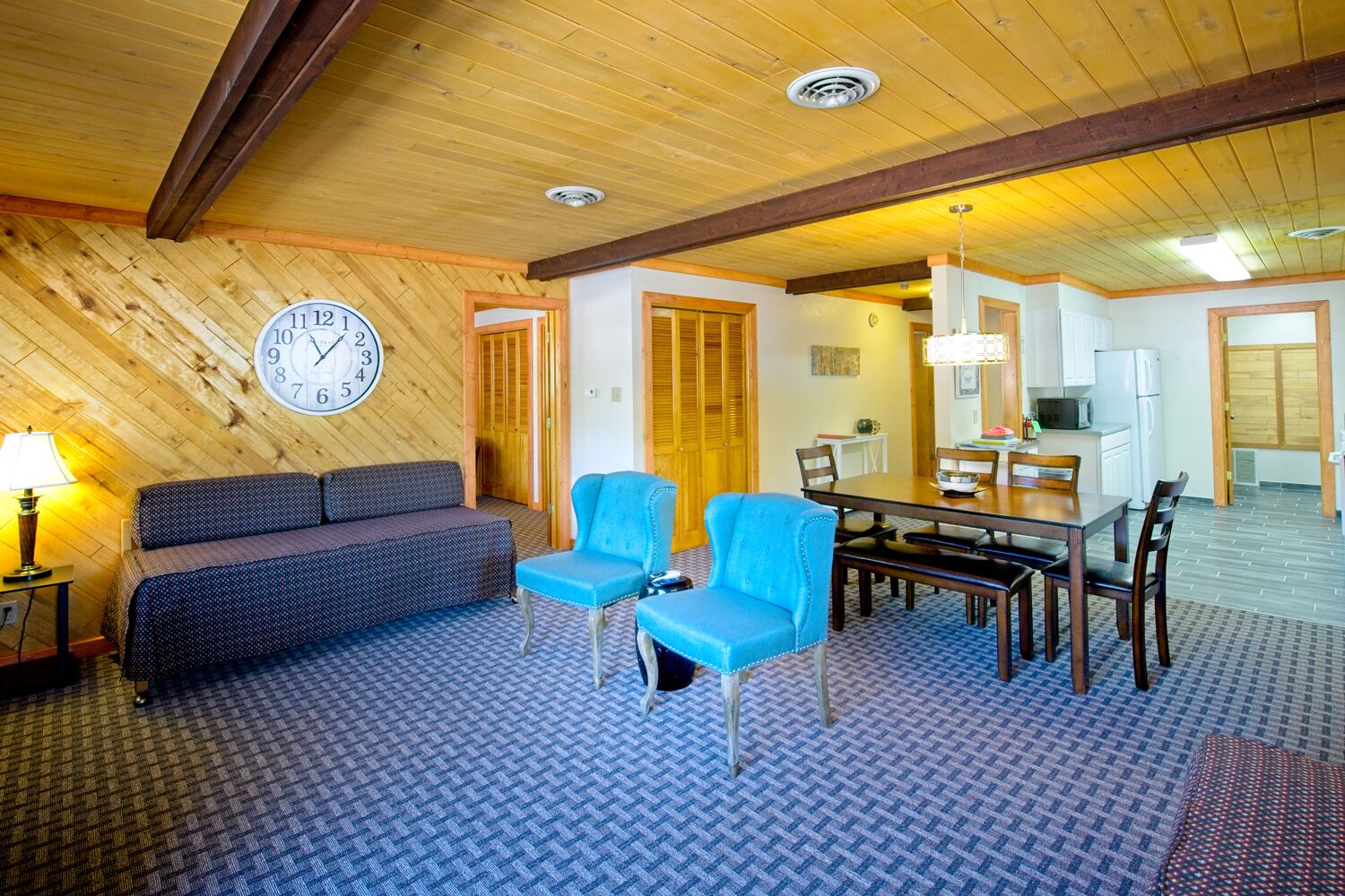 A cozy room with a wooden ceiling, a clock, a sofa, two blue chairs, a dining table with chairs, and a kitchen area.