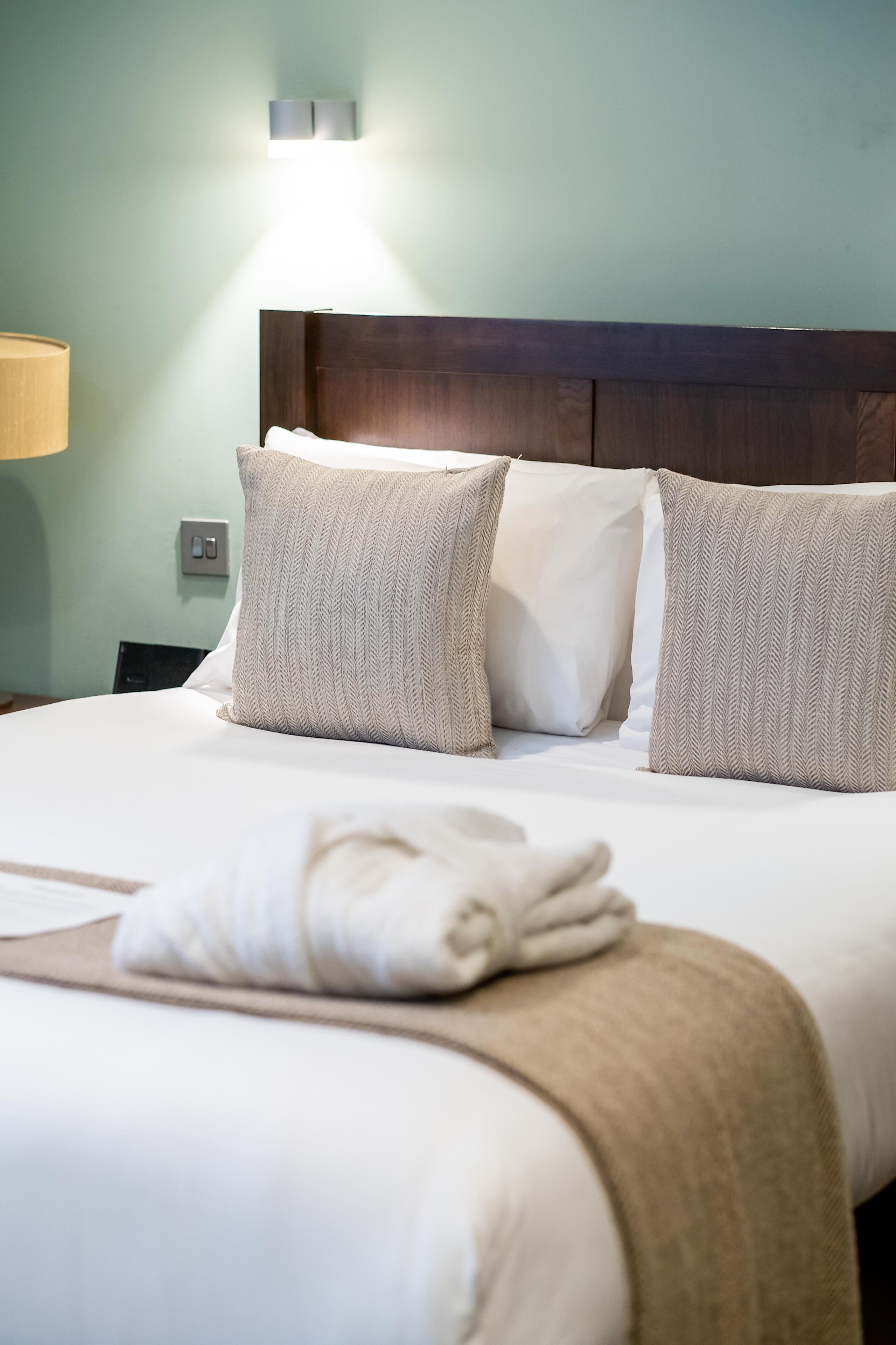 A neatly made bed with white linens and beige pillows, a wooden headboard, and a wall-mounted light fixture.
