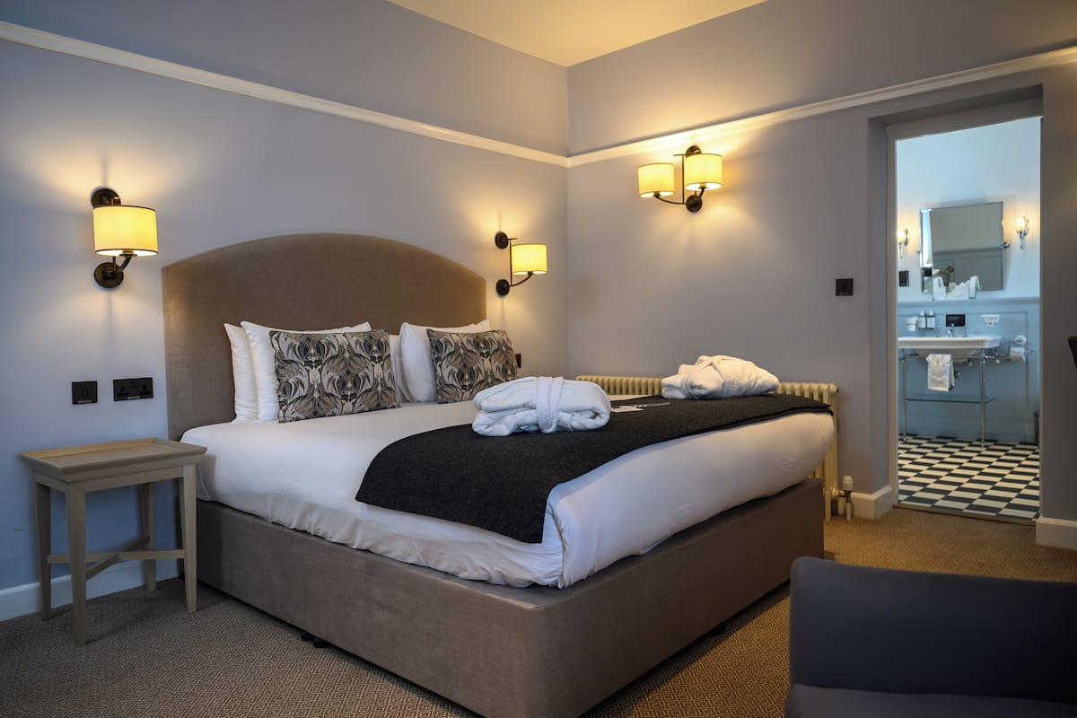 Habitación de hotel con una cama grande, lámparas de noche, una mesita y vista al baño.