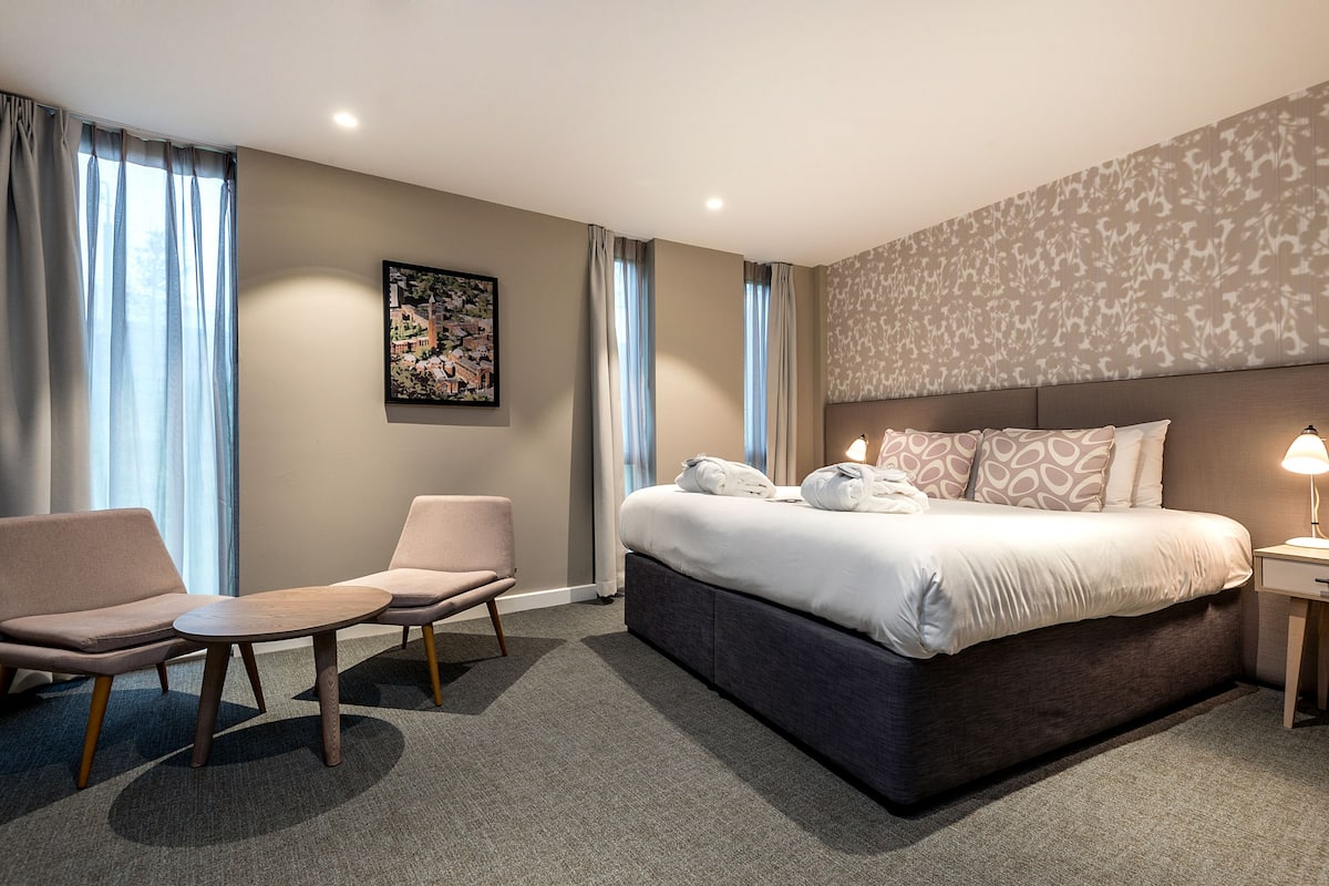 Una habitación de hotel moderna con una cama grande, dos sillones, una mesita y una mesita de noche con lámpara.